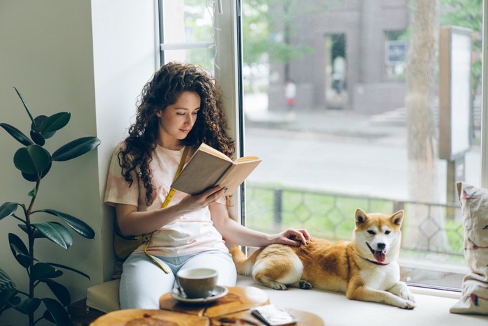 窓辺で本を読む女性と隣に座る柴犬